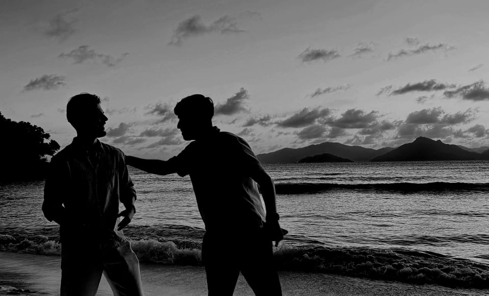 Zwei Männer in hellen Hemden stehen bei Sonnenuntergang an einem Strand, im Hintergrund Meer und Inseln. Schwarzweiß.
